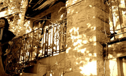 Photograph of a woman standing on the steps of an old house. The parts of the house are falling apart, and the area is overgrown with plants.