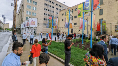 A lively outdoor event with attendees standing near colorful banners and modern architecture on a cloudy day, surrounded by greenery.