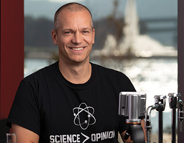 A portrait of Jason Brown sitting in a wet lab with a t-sheet with type saying science is greater than opinion.