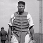 A black and white photo of an Black baseball player wearing a catchers vest.