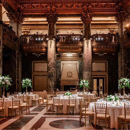 Elegant dining hall with ornate decor, featuring round tables set with floral centerpieces and golden chairs under grand architecture.