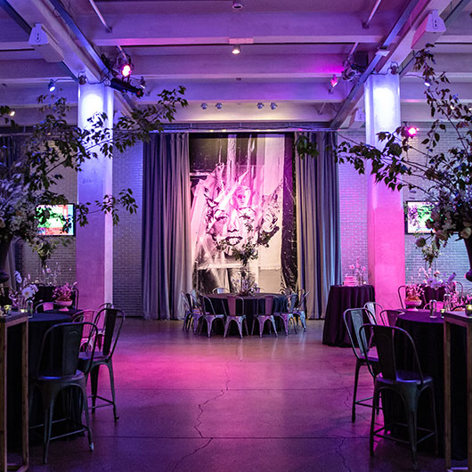 Elegant event space featuring purple lighting, chic tables, and a large artistic backdrop surrounded by greenery.