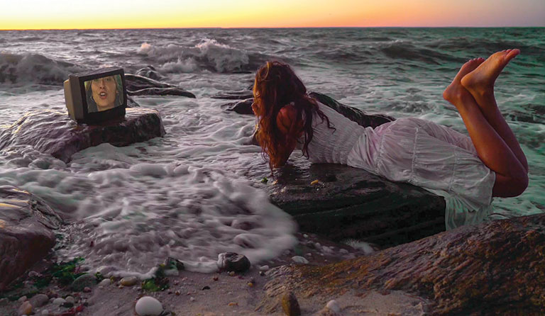A video clip of a young woman laying on rocks at a beach staring at a small tv