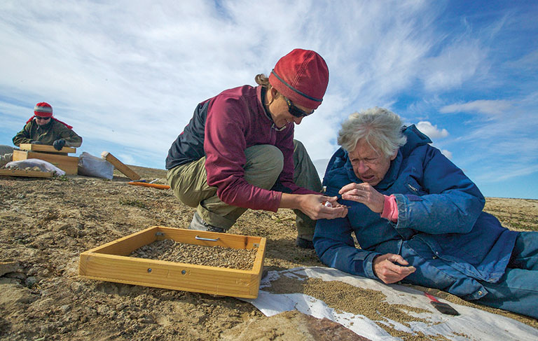 2 paleontologists kneeling and laying on the ground inspecting some fossil finds