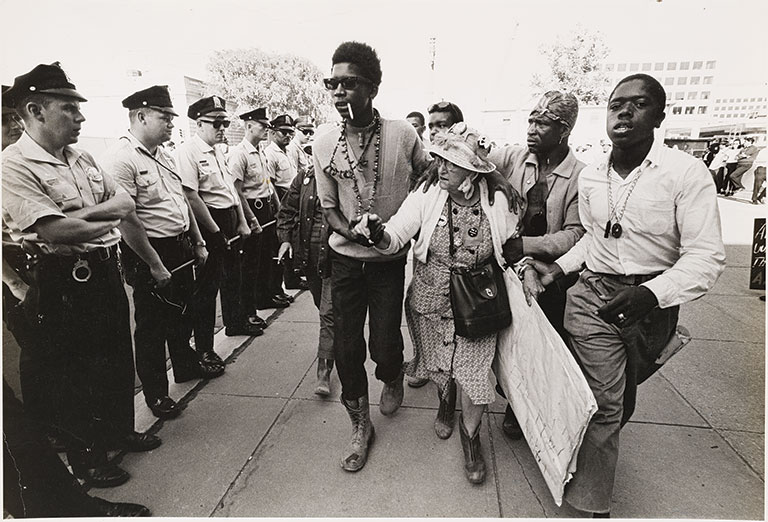 A group of men helping an elderly african american women past a line of policemen
