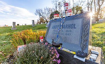 A tombstone with the name Andy Warhol, with campbell soup cans and bananas placed on it.
