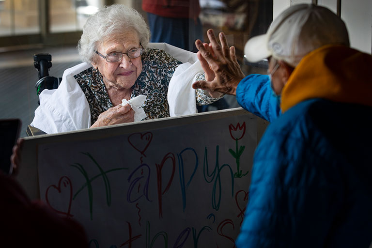 An elderly woman touching her son's hand through a glass partition.