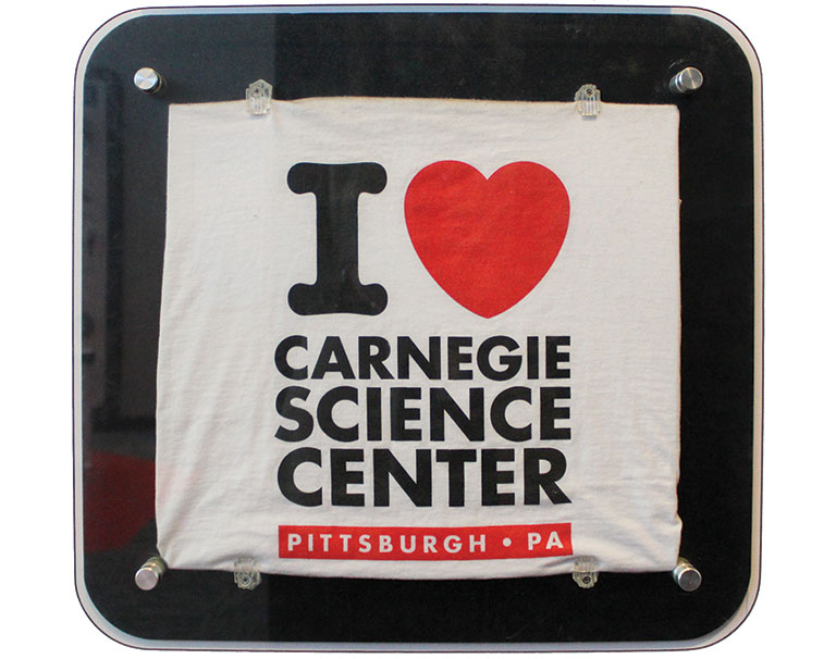 A t-shirt with the words I heart Carnegie SCience center, framed in a display case.