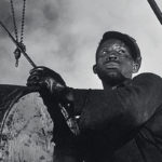 a man looks into the distance while handling a large vat of grease.