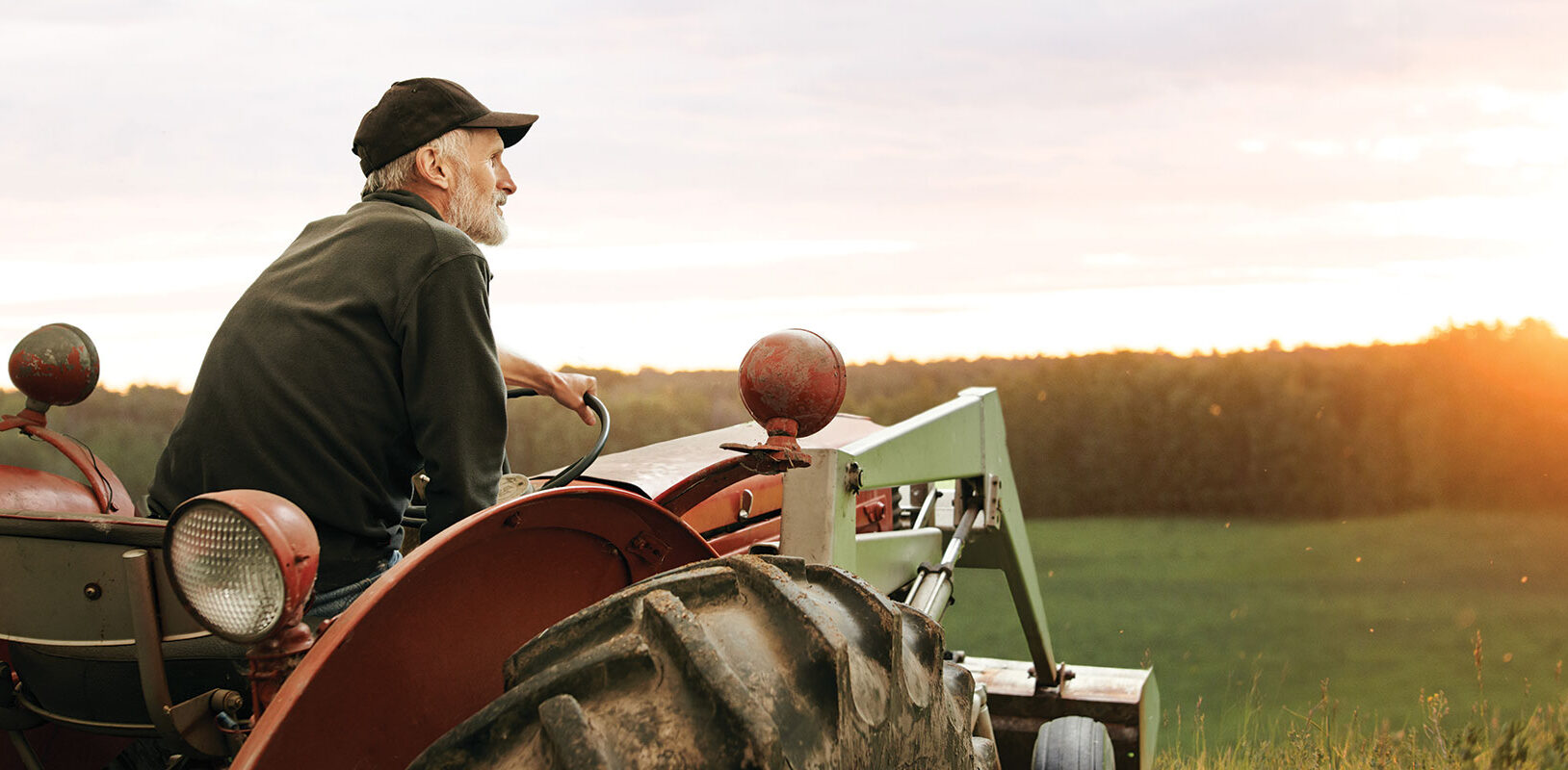 A farmer sittin gon a tractor looking over a field during sunset.