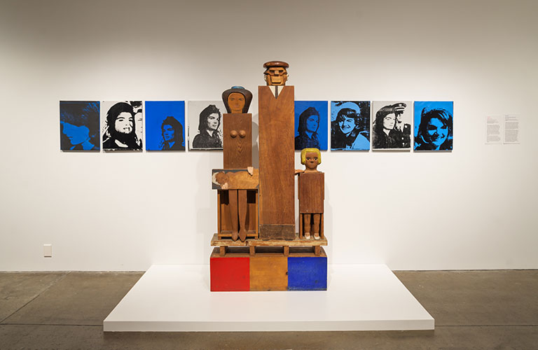 An art installation of a family of wooden boxy figures in front of a wall of painting of jackie kennedy