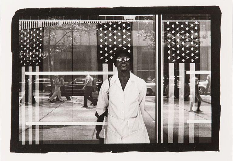 Black and white photo of a man standing in front of a window that's reflecting the american flag