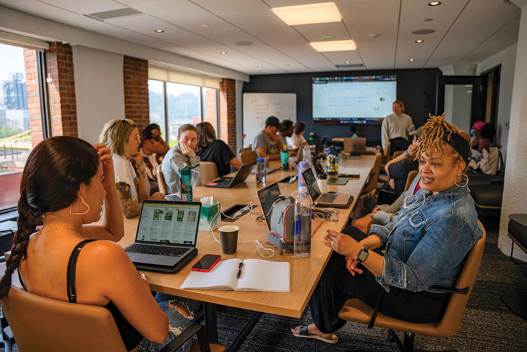 A group of adult students working on laptops while sitting around a large table.