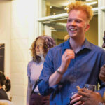 A group of three people engage in conversation at a social event, smiling and laughing, with others in the background.