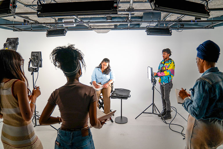 A group of four diverse individuals in a studio observes a woman sitting on a chair, preparing for a shoot.